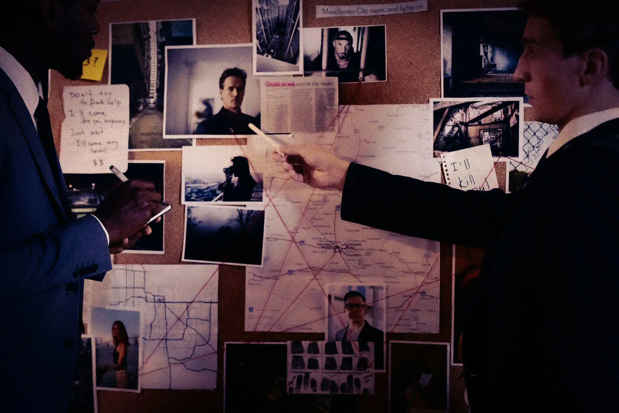 Two detectives in suits study a cork board covered with photographs, notes and a map connected by red string.