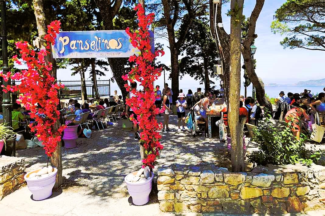 Eingang des Biergartens Panselinos auf Alonnisos mit pinkfarbenen Blumen vielen Menschen zwischen Bäumen