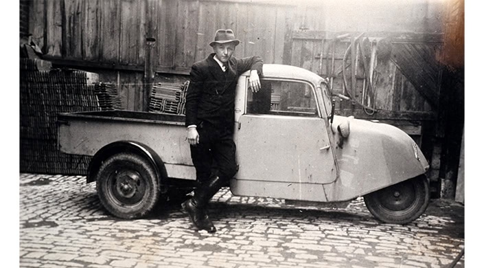 Ein Mann in Anzug und Hut lehnt an einem Auto, schwarz-weißes Foto auf einem Kopfsteinpflaster, Hintergrund mit Holzwand.