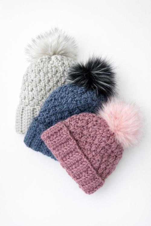 Three beanies with pom-poms—gray with white, blue with black, and pink with light pink—are displayed on a white background. Free crochet pattern