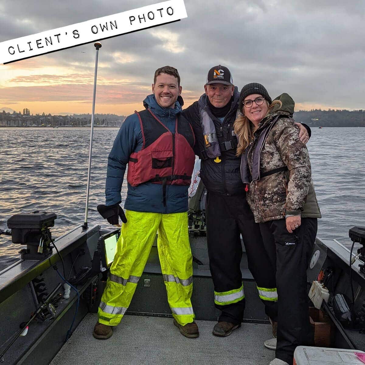 Three people in outdoor gear stand on a fishing boat at sunrise, smiling at the camera. Water and a distant shoreline are visible in the background.