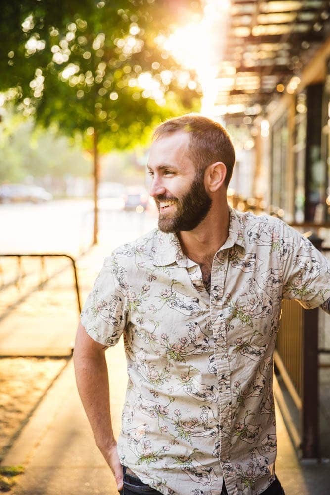 A man with a beard smiles while standing outside on a sunny day, wearing a patterned short-sleeve shirt. Sunlight filters through trees and a metal awning in the background.