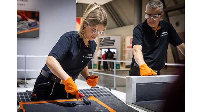 Eine Frau mit Schutzbrille und orangefarbenen Handschuhen arbeitet an einem technischen Projekt auf einem Tisch, im Hintergrund sind weitere Personen und Ausstellungsstücke zu sehen.