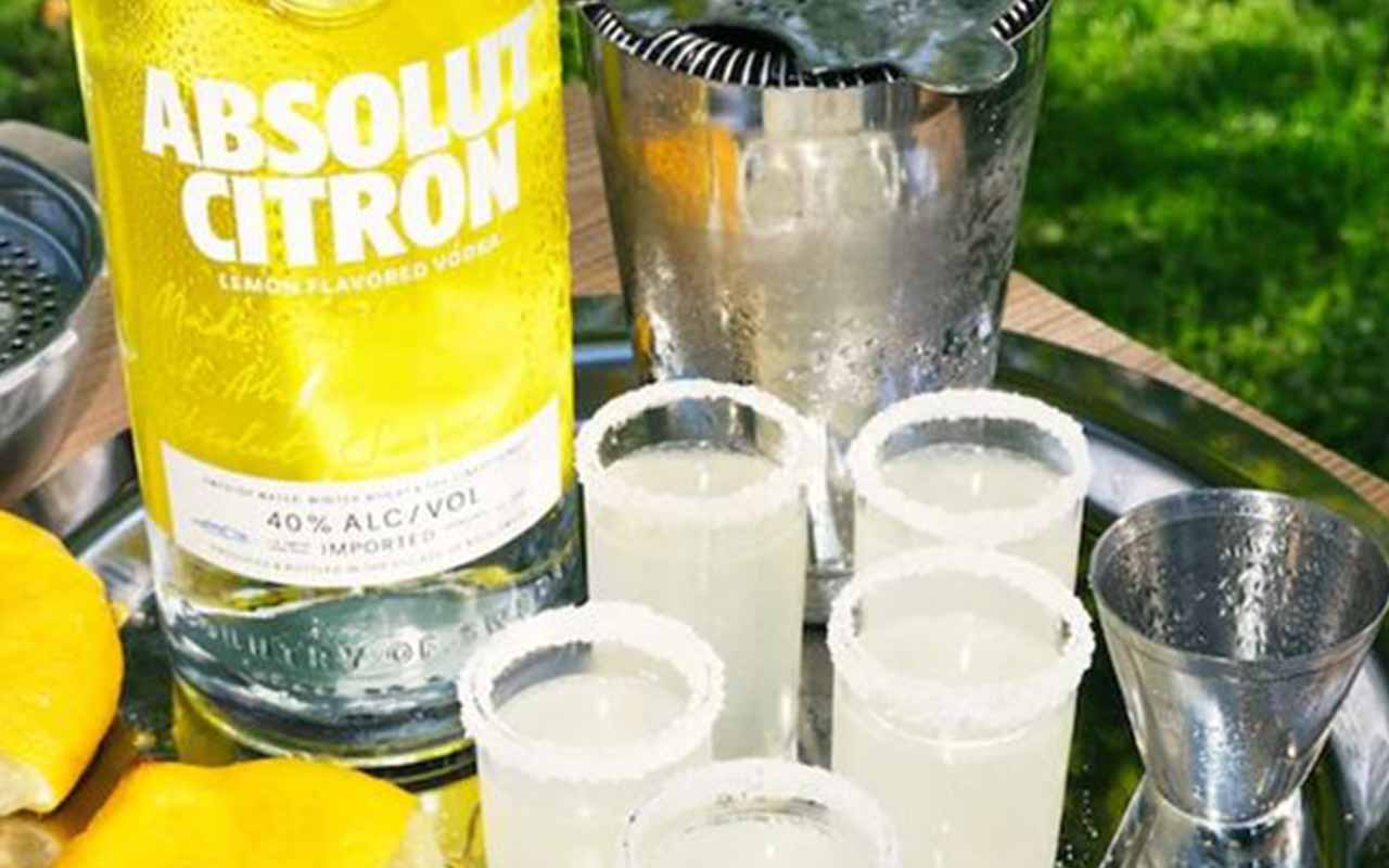 A bottle of Absolut Citron vodka, four salt-rimmed shot glasses filled with a clear drink, a jigger, and lemon slices on a metal tray outdoors.