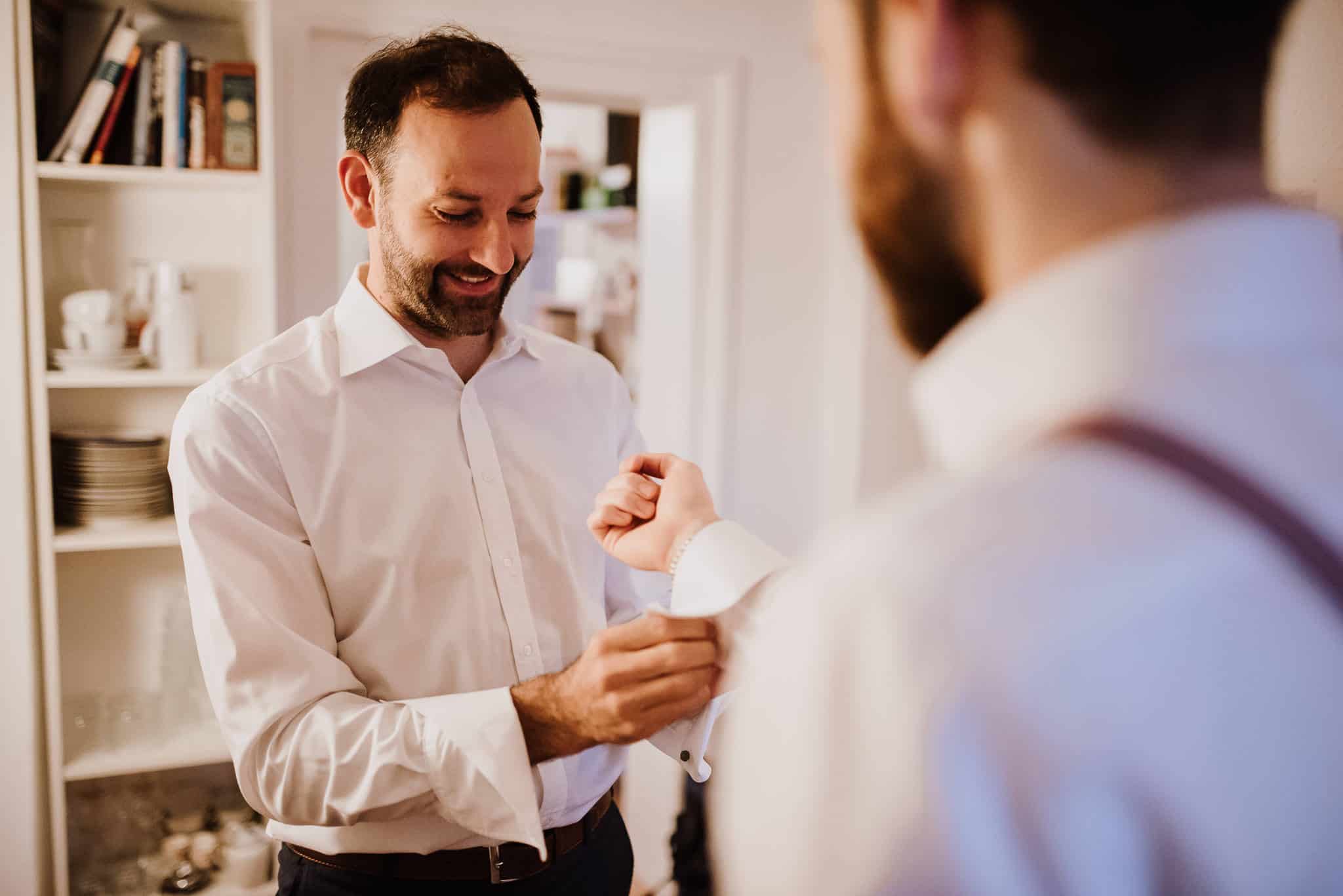 Zwei Männer lachen beim Anlegen der Manschettenknöpfe – echte Hochzeitsmomente in NRW.