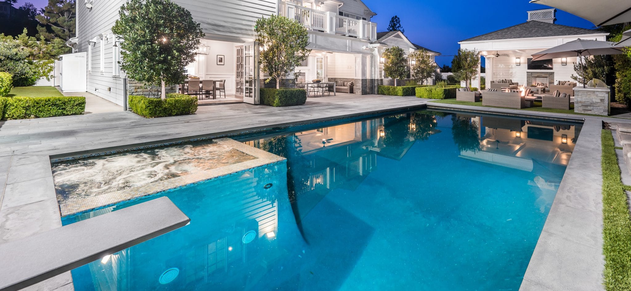 Modern house with illuminated windows, outdoor patio seating, and a large swimming pool with attached spa in the backyard during dusk.