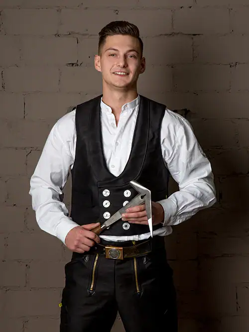 Dachdeckermeister Maik Kötter in traditioneller Zunftkleidung hält ein Zimmermannswinkel vor einer gemauerten Wand stehend.
