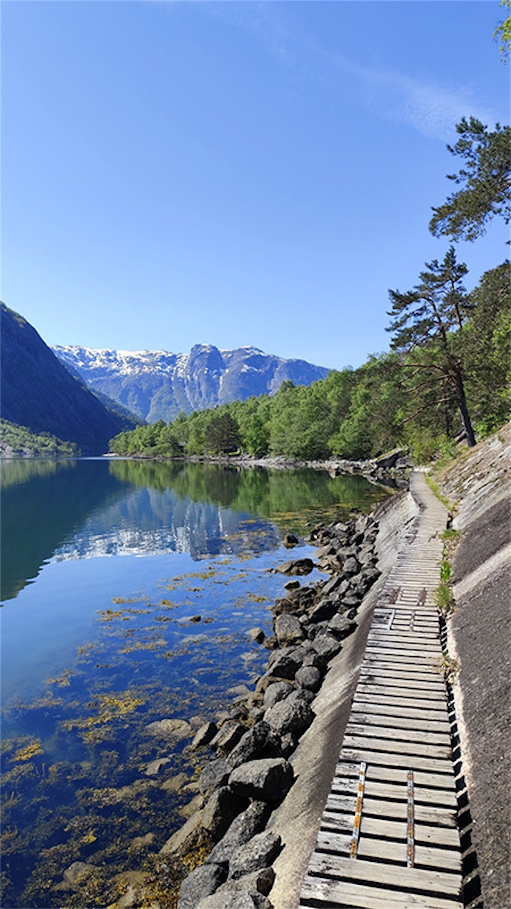 Klarer See mit Felsen im Vordergrund, bewaldete Ufer und schneebedeckte Berge im Hintergrund unter blauem Himmel.