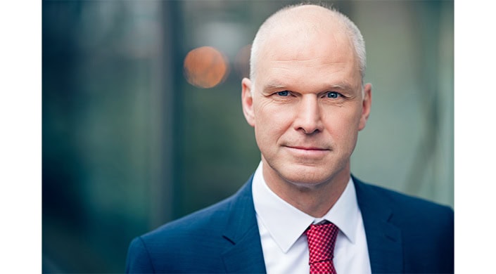 Nahaufnahme eines Mannes im blauen Anzug mit weißem Hemd, nur die rechte Gesichtshälfte sichtbar, verschwommener Hintergrund.
