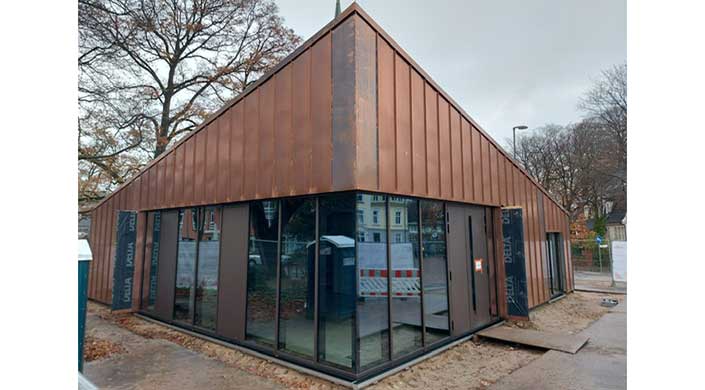 Eckiges Gebäude mit Glasfront und Kupferverkleidung, im Hintergrund Bauabsperrung und mobile Toiletten, spiegeln sich in den Fenstern.