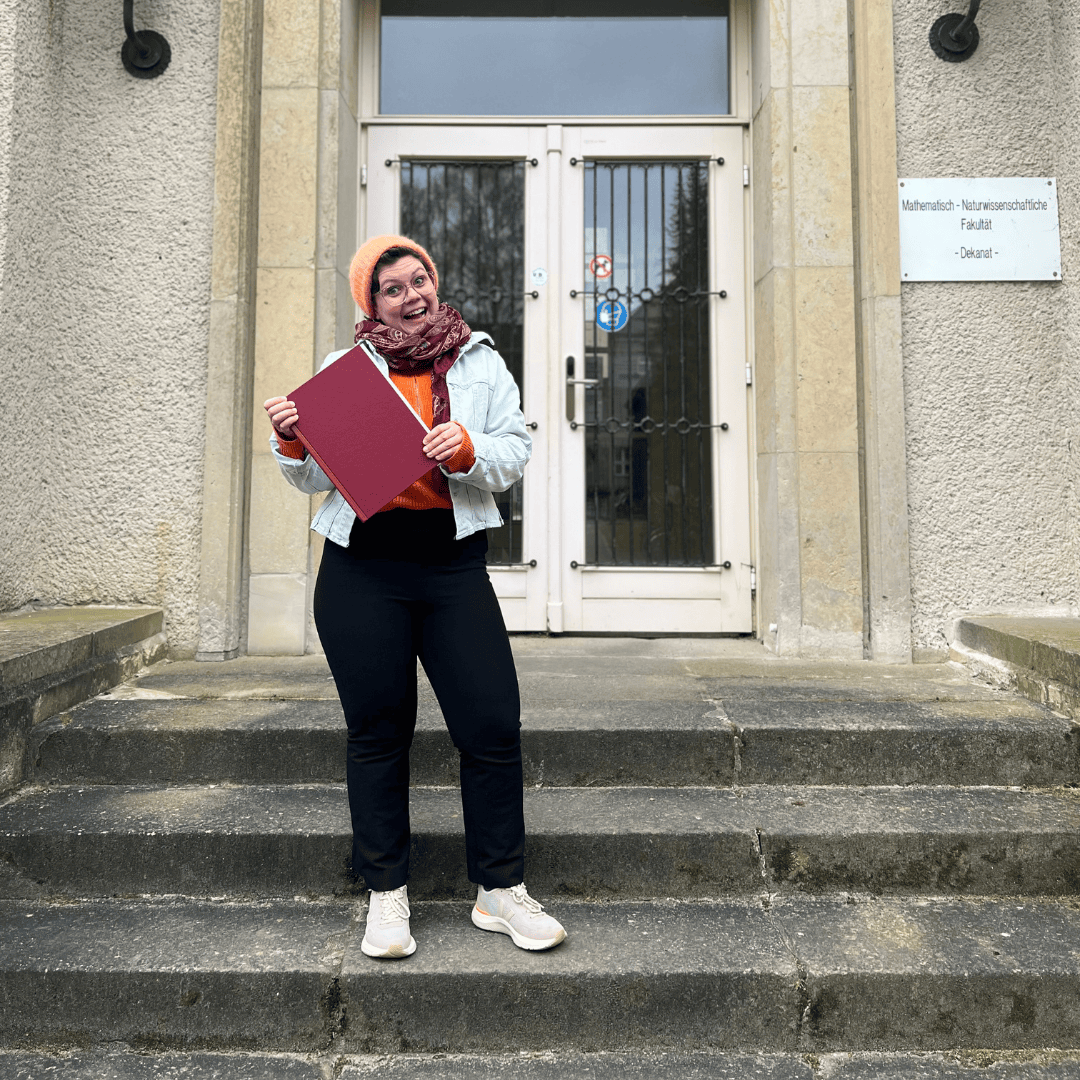 Cosma_Hoffmann Im Mittelpunkt des Bildes steht eine fröhlich lachende Frau – Cosma Hoffmann – die auf den Stufen eines Gebäudes steht. Sie hält ihre Dissertation in der Hand. Ihr Gesichtsausdruck vermittelt Stolz, Erleichterung und Freude.