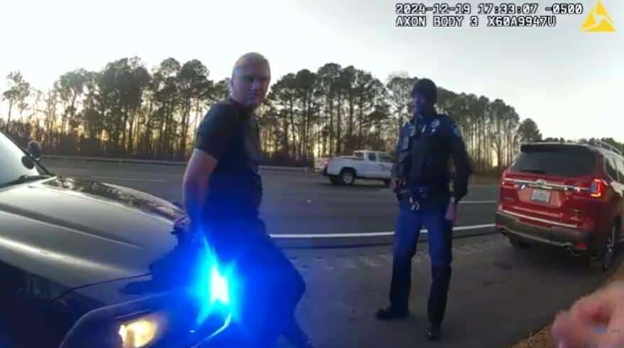 A man in handcuffs is detained by a police officer on a highway, with a police car's blue lights illuminating the scene. A red SUV and a white pickup truck are visible in the background. This could be related to a Florida doctor arrest case.