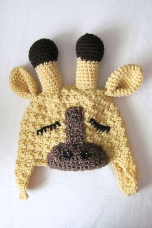 Crocheted yellow giraffe hat with brown horns, ears, snout, black nostrils and eyelashes, laid flat on a white surface. Free crochet pattern