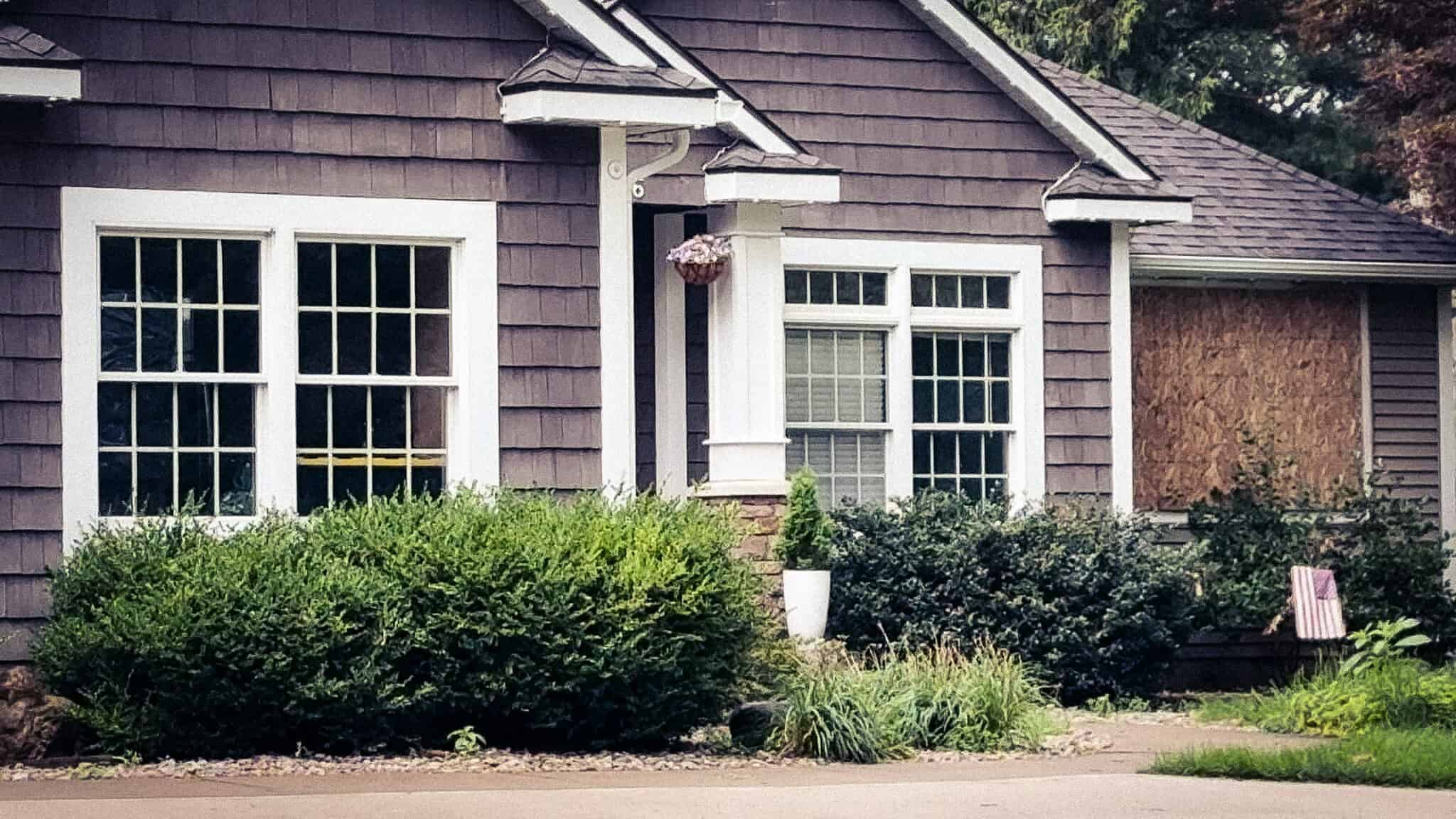A grey shingled suburban house with white-framed windows and shrubs along the front garden.