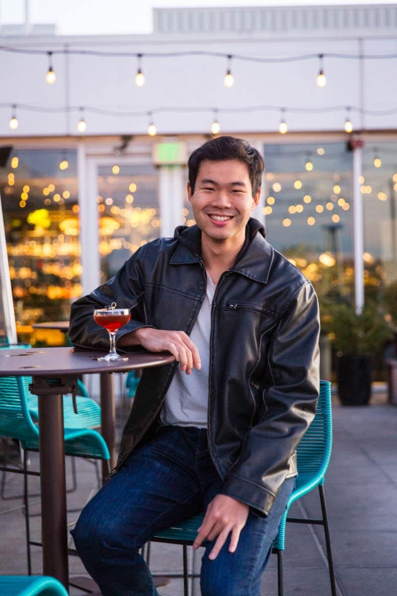 A person in a leather jacket sits at an outdoor table with a cocktail. String lights are visible in the background.
