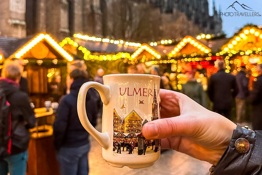 Eine Tasse des Ulmer Weihnachtsmarktes mit beleuchteten Ständen im Hintergrund