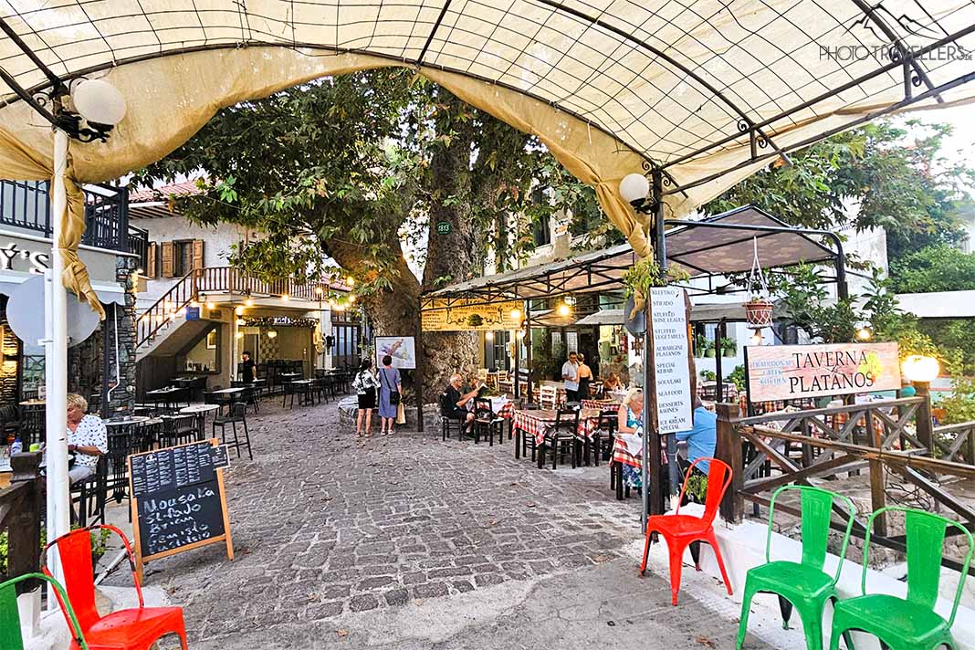 Grüne und rote Stühle auf Pflastersteinen unter einem Stoffdach mit einer gut besuchten Taverne dahinter