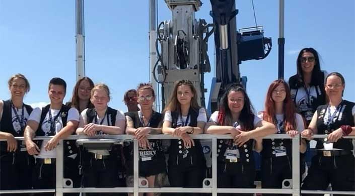 Zwei Frauen stehen draußen vor einer großen metallischen Maschine, tragen Namensschilder und Westen, bei sonnigem Wetter.