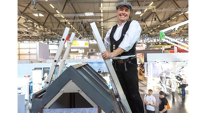 Ein Handwerker mit traditioneller Kleidung hält eine Metallleiste, steht neben einem Dachmodell auf einer Messe oder Ausstellung.