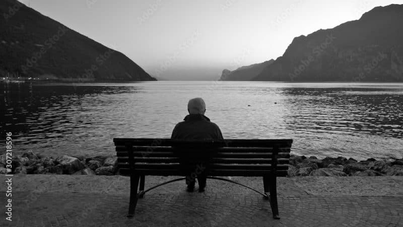 A lonely old man sits on a wooden bench at dusk after sunset, looking at the lake and the light on the horizon.