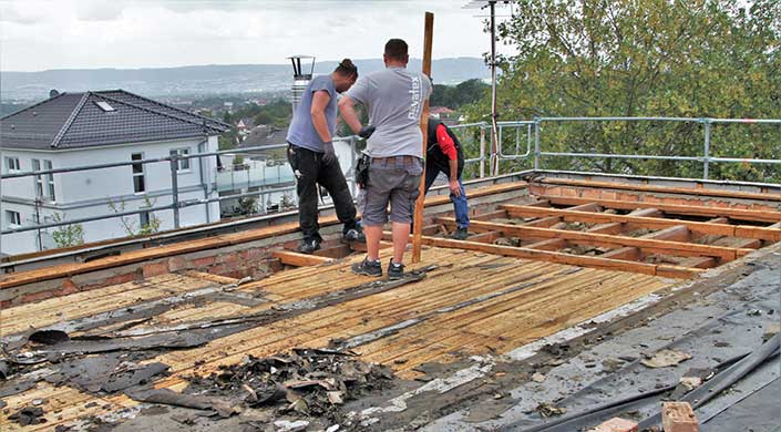 Drei Männer arbeiten auf einer Baustelle im Freien, tragen legere Kleidung und stehen auf einem teilweise abgedeckten Holzgerüst.