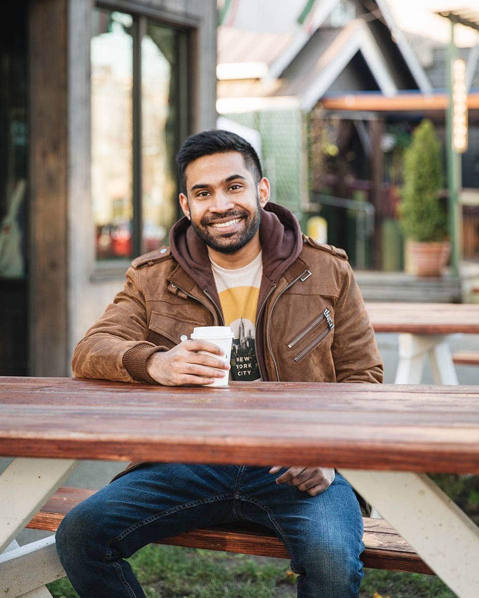 A man sits at a wooden picnic table outdoors, smiling at the camera and holding a takeaway coffee cup.