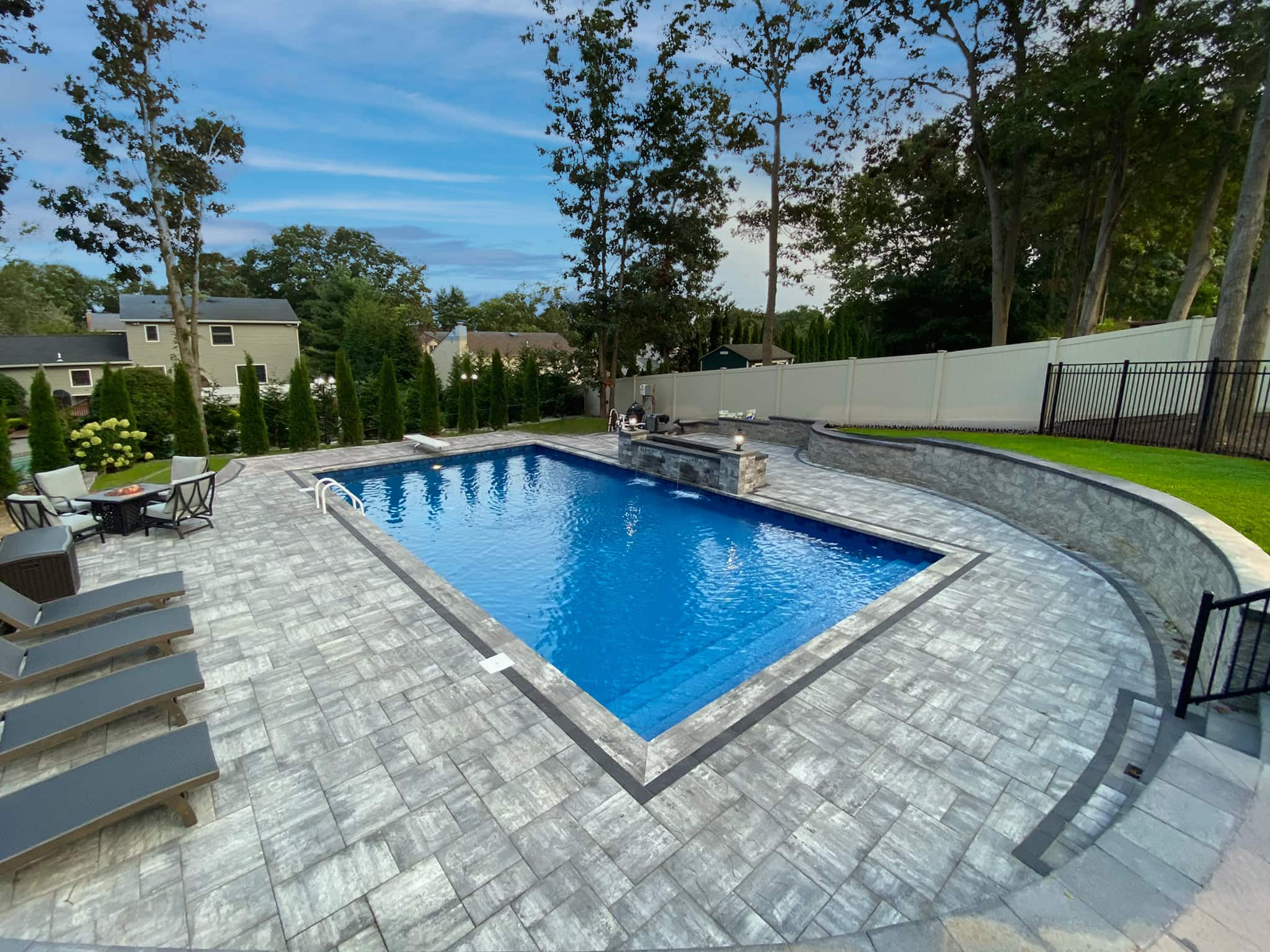 Rectangular in-ground swimming pool with a vinyl pool renovation, surrounded by gray stone patio, lounge chairs, dining area, and a landscaped backyard with trees and privacy fencing.