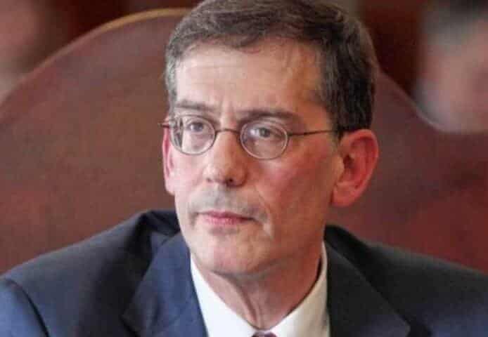 A middle-aged man with graying hair and glasses, wearing a dark suit and white shirt, looks intently at something off-camera. He appears to be in a professional setting.