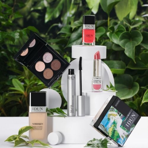 Een compositie van IDUN Minerals make-upproducten tentoongesteld op witte, cirkelvormige blokken tegen een achtergrond van groene bladeren. De collectie bevat een foundationfles, een open oogschaduwpalette met neutrale tinten, een mascara met een zilveren huls, een rode nagellak, een roze lipstick en een compact poeder met een artistieke verpakking. De natuurlijke elementen en frisse kleuren benadrukken de pure en minerale samenstelling van de producten.