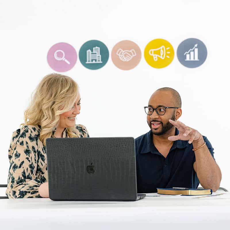 A marketing agency professional collaborates with a client at a laptop under strategy icons.