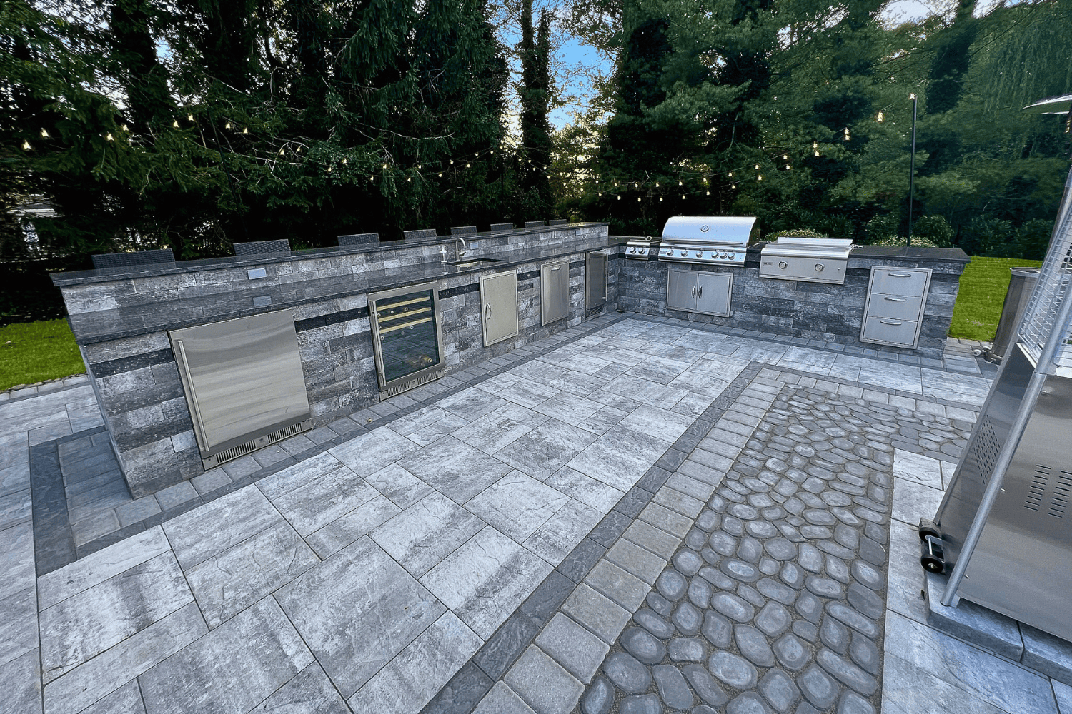 Outdoor Kitchens feature stainless steel appliances, gray stone counters, and tiled flooring set in a landscaped backyard with trees and string lights overhead.