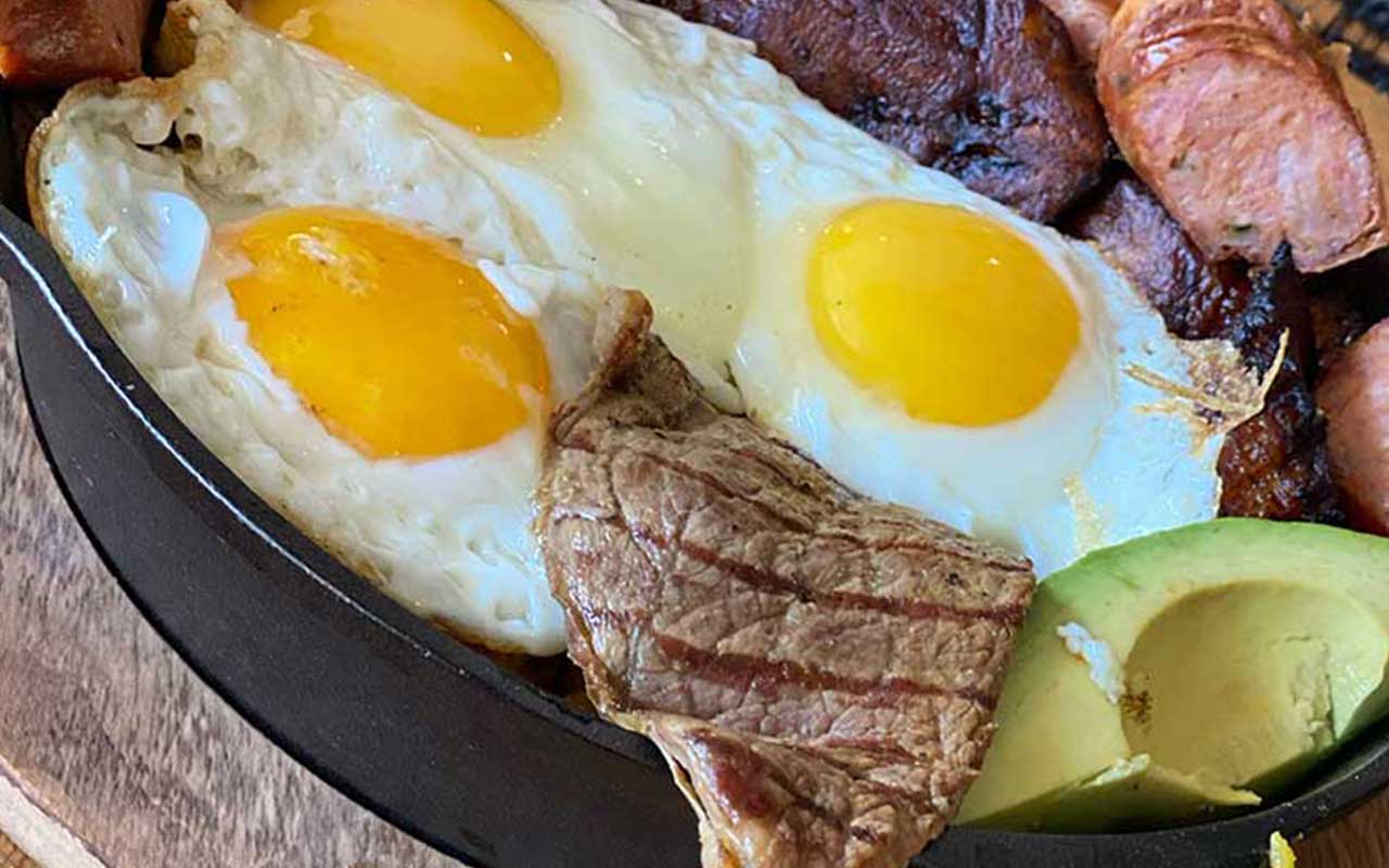 A skillet with two sunny-side-up eggs, grilled steak, sliced sausages, and avocado.