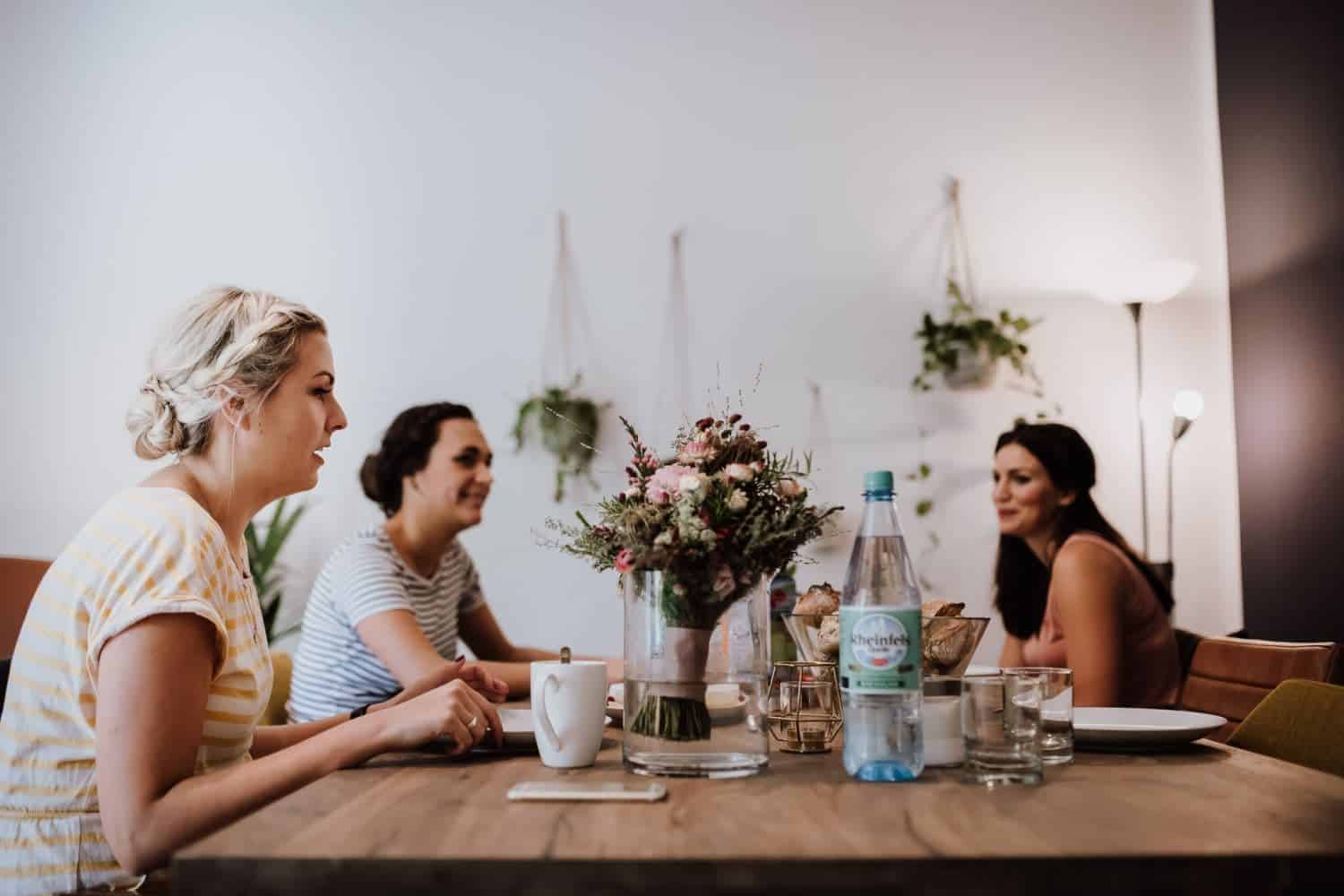 Drei Frauen lachen an einem hellen Holztisch beim Kaffee – echte Momente für Hochzeitsreportage NRW.
