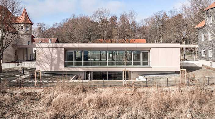 Modernes Gebäude mit großen Glasfenstern, umgeben von trockener Wiese und Bäumen im Hintergrund.