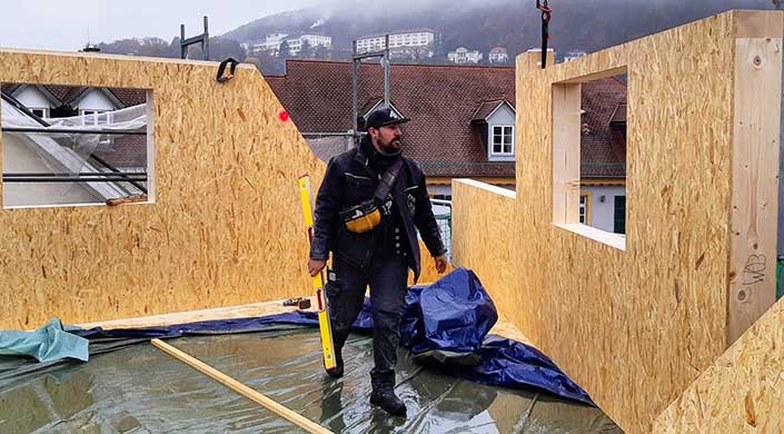 Ein Bauarbeiter in schwarzer Kleidung und Mütze trägt ein Messgerät auf einer Baustelle im Freien. Im Hintergrund sind Häuser und Berge zu sehen.
