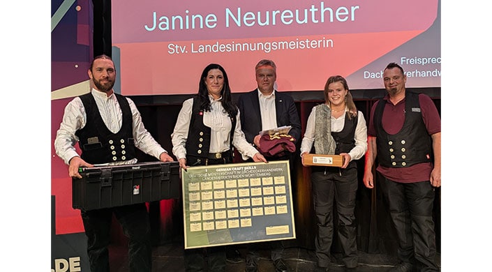 Bild von Landessiegerin Janine Neureuther Zwei Personen stehen auf einer Bühne, halten eine gerahmte Tafel und eine rote Stoffjacke. Im Hintergrund ist ein Bildschirm mit Text.