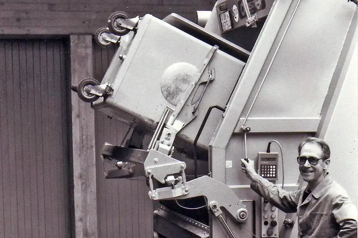 A smiling man is opening the tailgate of a garbage truck
