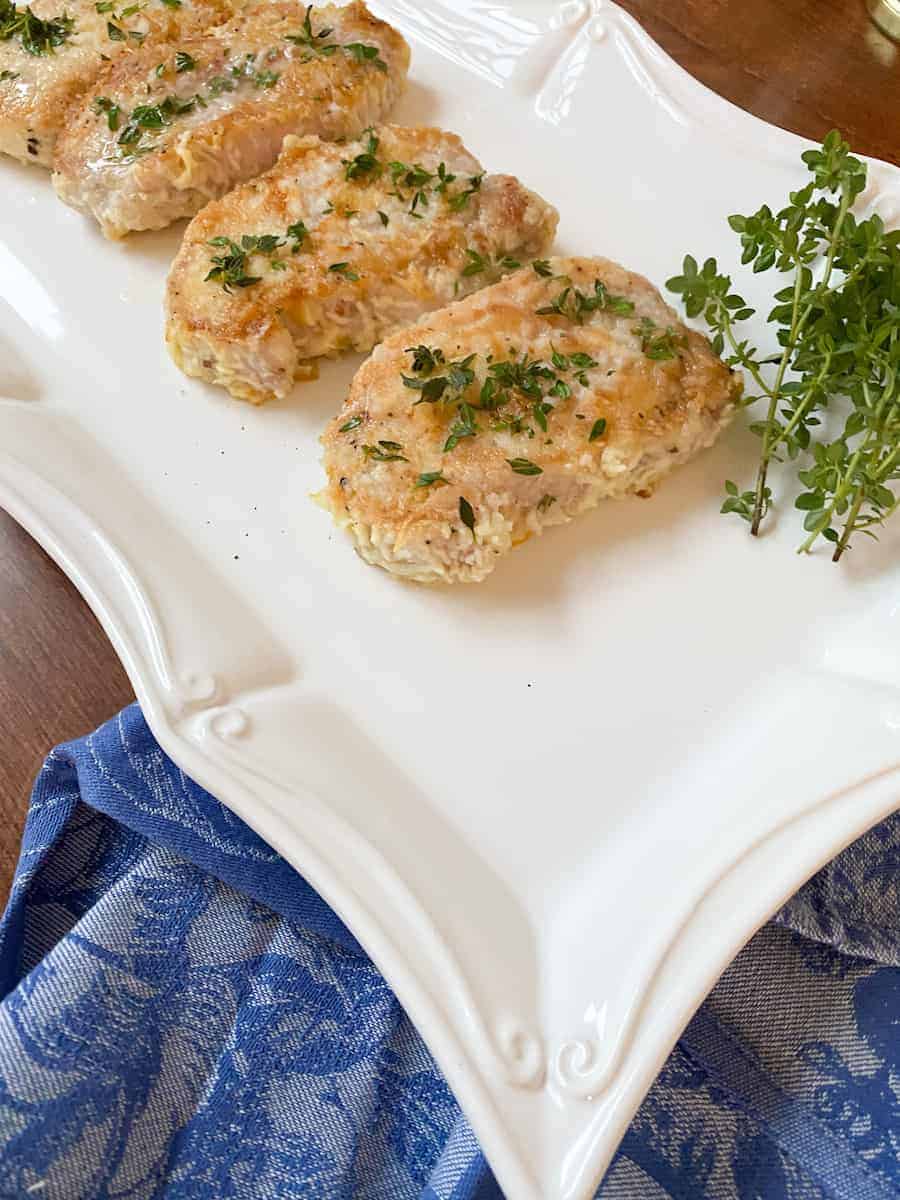 gluten-free cheese-crusted pork chops plated on a white platter with fresh thyme garnish. There is a blue tea towel on the side