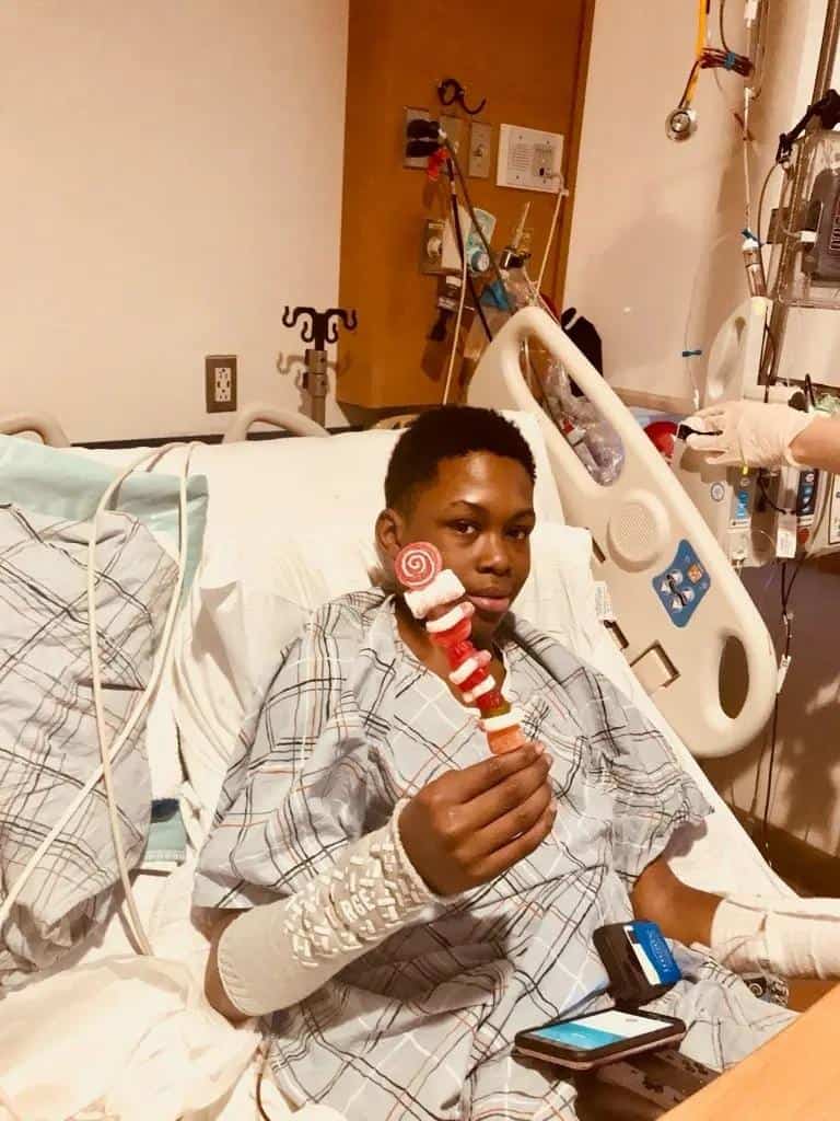 Young Black patient in a hospital bed, smiling and holding a candy kebab skewer, likely after receiving a "sickle cell disease" treatment. The setting is a hospital room, suggesting recovery and "pain relief.