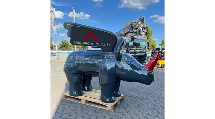 Große, glänzende Statue eines stilisierten Tieres mit Flügeln, steht auf einer Holzpalette im Freien unter blauem Himmel.