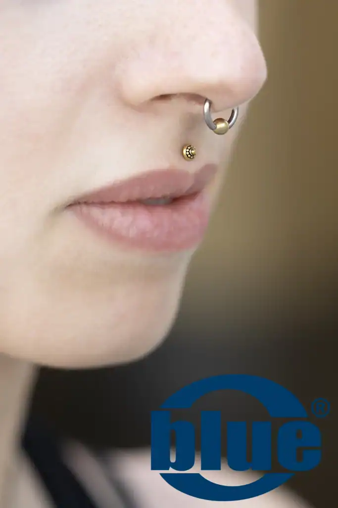 Close-up of a person's face showing a silver septum hoop with a gold bead and a small gold cheek stud, with a blue logo partially visible at the bottom right.