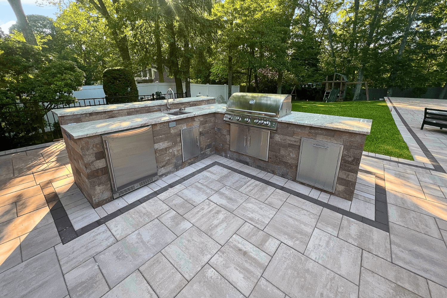 Outdoor Kitchens feature stone countertops, a built-in grill, sink, refrigerator, and storage cabinets, all set on a tiled patio surrounded by trees and lawn.