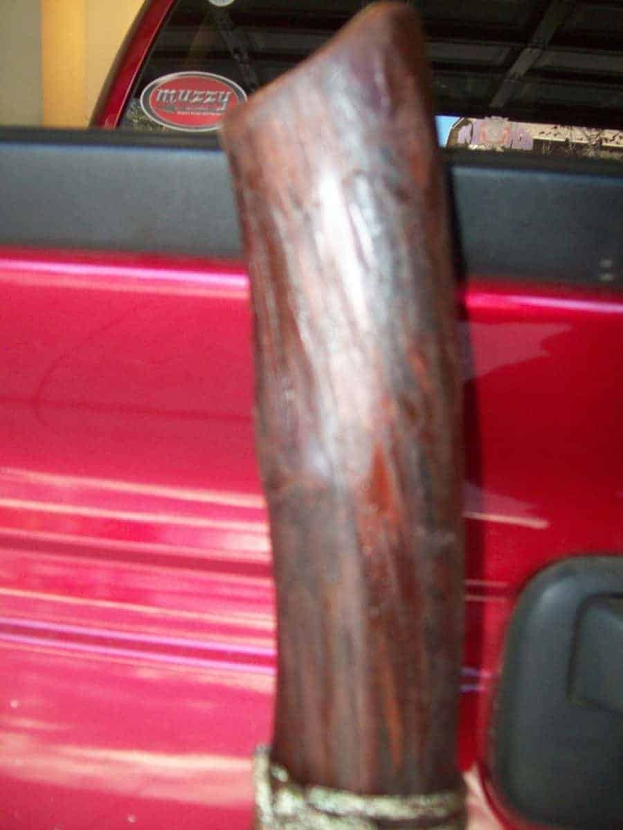A close-up of a handcrafted, lacquered wooden walking stick, likely an oak branch staff, with a rich mahogany finish. The staff is hand wrapped with twine near the base, emphasizing the handmade staff process. The red background suggests it may be resting against a red vehicle, perhaps a custom item from wood.