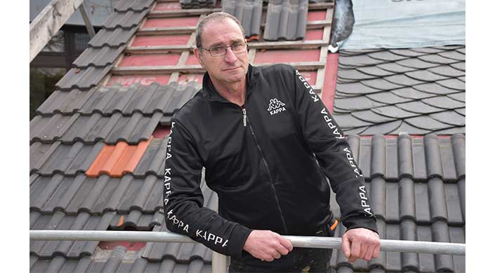 Ein Mann mit Brille trägt eine schwarze Kappa-Jacke und lehnt an einem Geländer vor einem Hintergrund mit Dachziegeln.