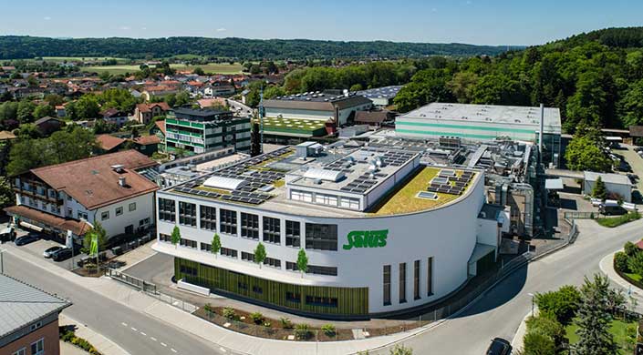 Modernes Firmengebäude mit dem grünen Schriftzug „SENNES“ an der Fassade und begrüntem Dach, umgeben von weiteren Gebäuden und einer grünen Hügellandschaft im Hintergrund.