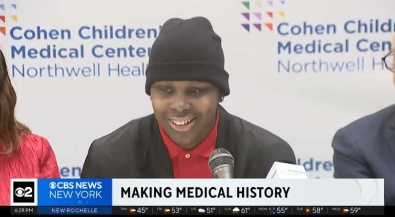 A smiling young Black man wearing a black beanie and red polo shirt speaks into a microphone at a press conference at Cohen Children's Medical Center. The background features the hospital's logo and the text "MAKING MEDICAL HISTORY," likely discussing a breakthrough treatment for sickle cell disease.