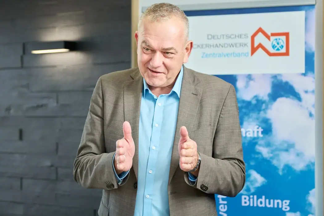 ZVDH-Präsident Dirk Bollwerk im grauen Anzug spricht mit Handgesten vor dem Hintergrund mit dem Logo des Zentralverbands des Deutschen Dachdeckerhandwerks.