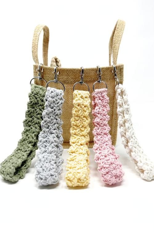 A jute bag with five crocheted keychains in green, gray, yellow, pink, and white—each made using a free crochet keychain pattern—hanging from the handle, displayed against a white background.