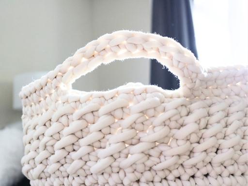 Close-up of a white woven basket with a handle, placed indoors near a window with sunlight highlighting its textured surface; no visible text or promotional information. Free crochet pattern