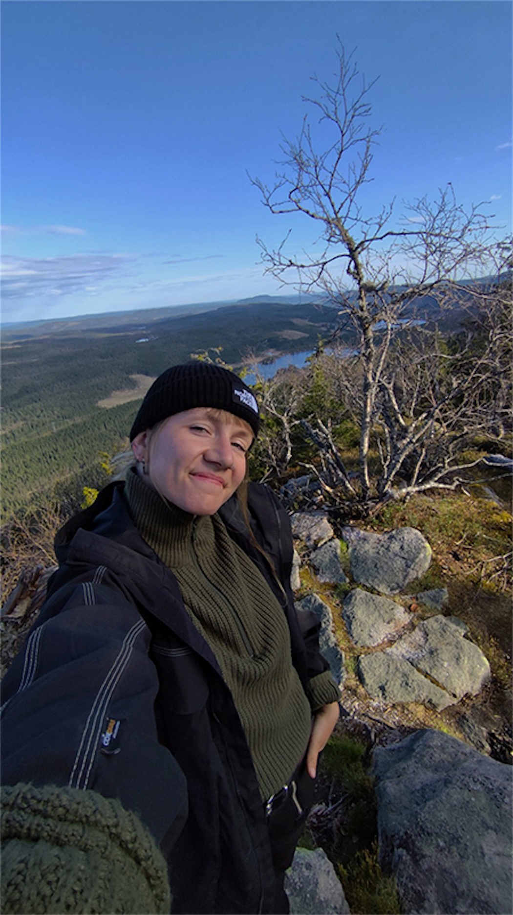 Frau mit Mütze macht Selfie in der Natur. Im Hintergrund Berge, Wasser und blauer Himmel.